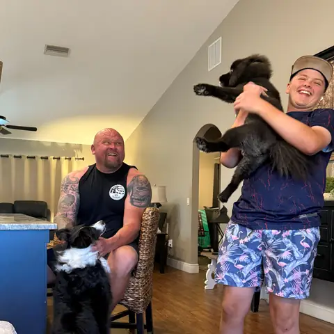 A young owner lifting his black dog at home while the trainer works with two others