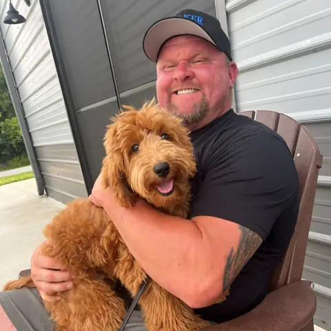 Chad Walker holding a full-grown red goldendoodle on his lap outside the training building