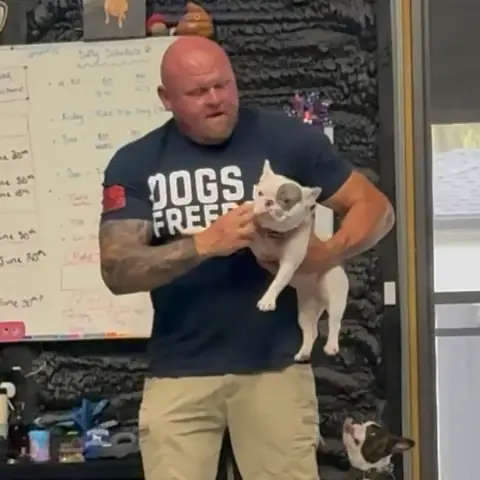 Chad Walker lifting a white French bulldog while a Boston terrier watches from the floor