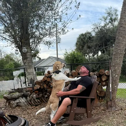 A tan dog up on its hind legs greeting its owner in a chair by the fire pit