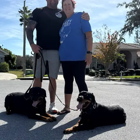 Chad Walker with a client in a blue shirt and her two black dogs holding a down-stay on the street